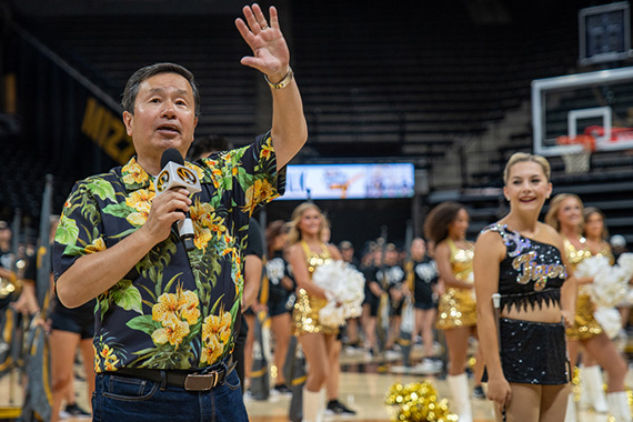Man with microphone speaking and waving from a basketball court