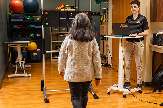 A person walking toward a machine while another person watches behind a computer