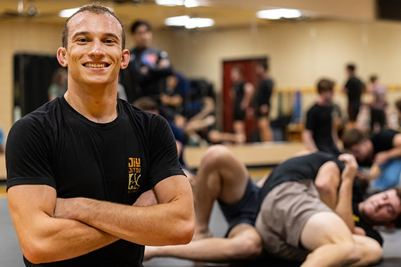 Student standing with arms crossed while others grapple on mat in background